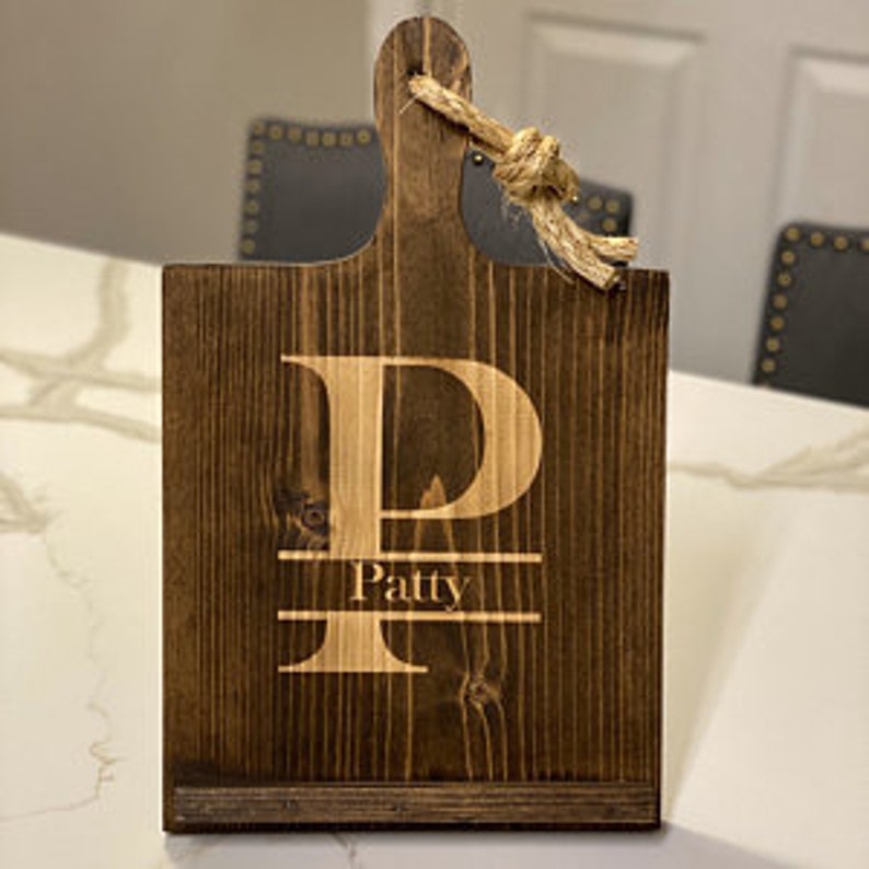 K&ouml;nnte beinhalten: Ein dunkelbraunes Holzbrett zum Schneiden mit einem gro&szlig;en wei&szlig;en Buchstaben "P" und dem Namen "Patty" eingraviert. Das Brett hat einen Griff mit einem geknoteten Seil.