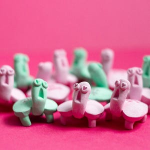 Puede incluir: Una colección de caprichosas figuras de tortugas hechas a mano en tonos rosa y verde menta. Las tortugas tienen caras expresivas con ojos en forma de corazón y bocas abiertas, sobre un fondo rosa vibrante. Estas figuras decorativas añaden un toque lúdico.