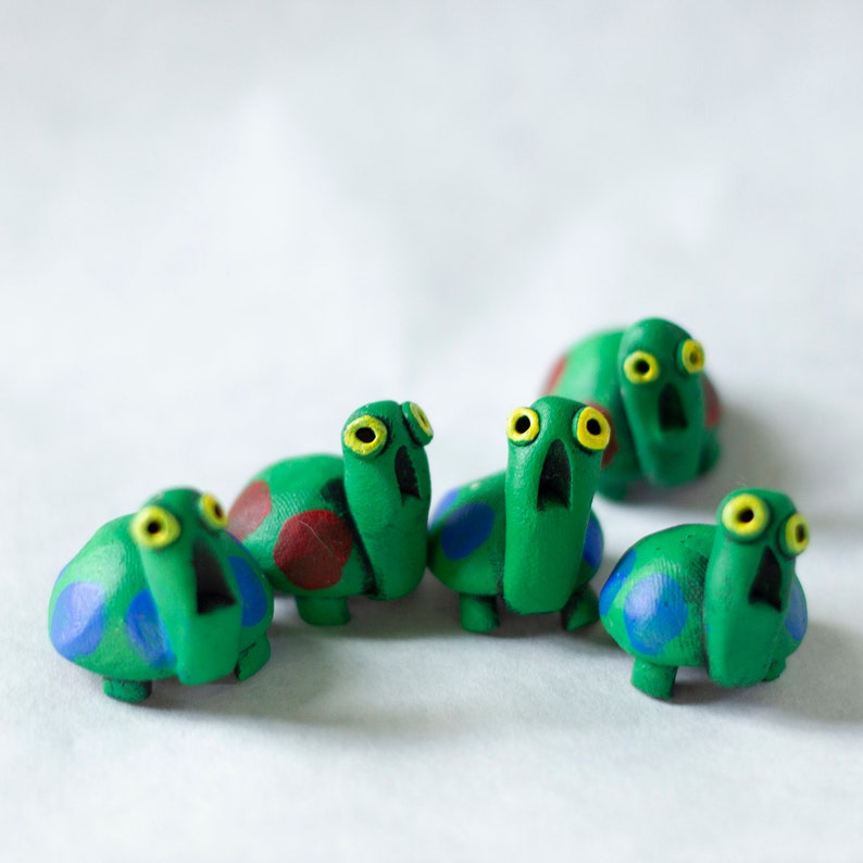 May include: A collection of six handmade, whimsical turtle figurines. Each turtle is painted bright green with yellow eyes, open mouths, and colorful shell details in red and blue. The turtles are arranged on a white surface.