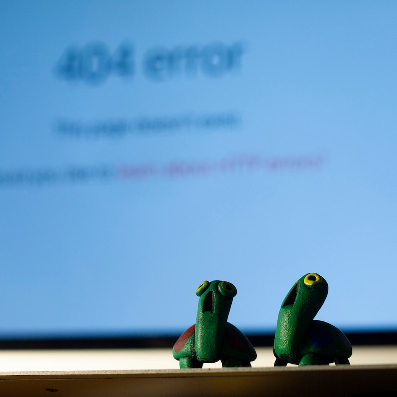 May include: Two green turtle figurines with yellow eyes and open mouths sit in front of a computer screen displaying a 404 error message. One turtle has a brown shell. The background is a light blue color.