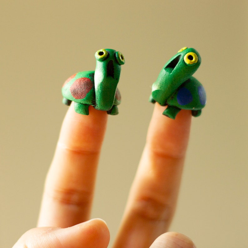 May include: Two handmade green turtle finger puppets with expressive eyes and colorful shell details. One turtle has red and brown spots, the other has blue spots. The puppets are displayed on fingers.