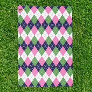 May include: A golf towel with a repeating argyle pattern in pink, green, white, and navy blue. The towel has a brass grommet and a loop for hanging. The towel is lying on a bed of green grass.