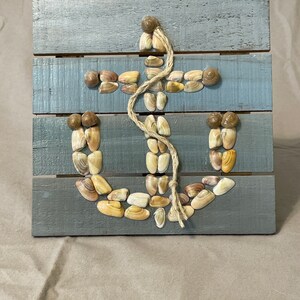 May include: A wooden sign with a blue background and a white anchor made of seashells. The anchor is decorated with a piece of twine.