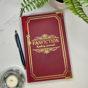 Peut inclure: Un journal rouge avec des accents dor&eacute;s et les mots "FANFICTION Reading Journal" sur la couverture. Le journal est ferm&eacute; et pos&eacute; &agrave; plat sur une surface blanche.