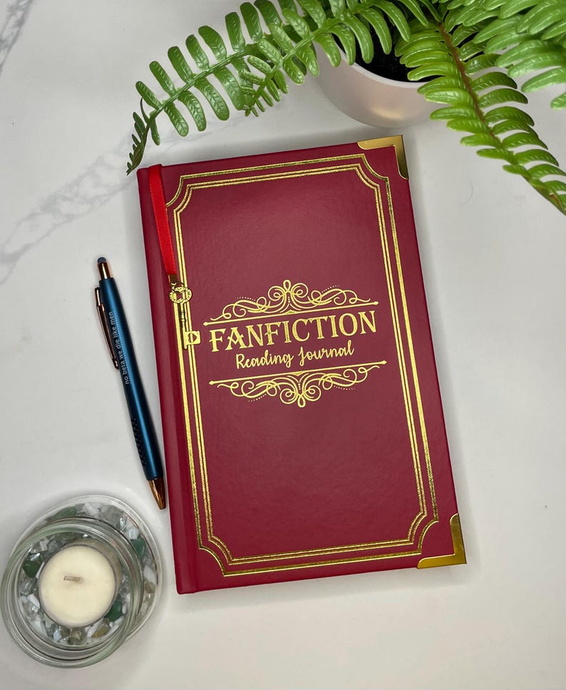 Peut inclure: Un journal rouge avec des accents dor&eacute;s et les mots "FANFICTION Reading Journal" sur la couverture. Le journal est ferm&eacute; et pos&eacute; &agrave; plat sur une surface blanche.