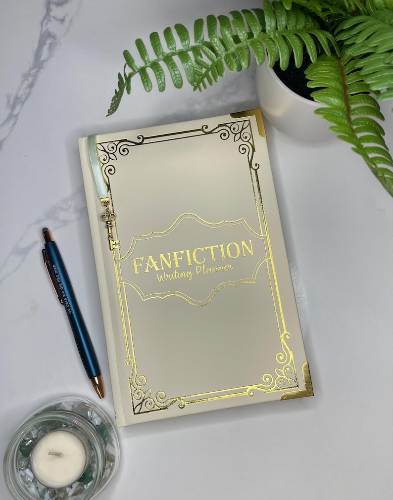 Pode incluir: Um caderno branco com uma borda dourada e as palavras "Fanfiction Writing Planner" na capa. Um chaveiro dourado em forma de chave pendura na parte superior do caderno.