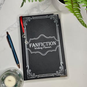 Pode incluir: Caderno preto com uma borda decorativa prateada e branca e o texto "FANFICTION Writing Planner" na capa. Um marcador de fita vermelha est&aacute; preso ao caderno.