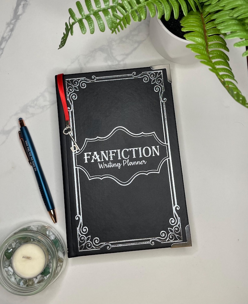 Pode incluir: Caderno preto com uma borda decorativa prateada e branca e o texto "FANFICTION Writing Planner" na capa. Um marcador de fita vermelha est&aacute; preso ao caderno.