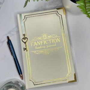 Peut inclure: Un journal blanc avec des accents dor&eacute;s et le texte "FANFICTION Reading Journal" sur la couverture. Un charme de cl&eacute; dor&eacute;e est attach&eacute; &agrave; un marque-page en ruban bleu.