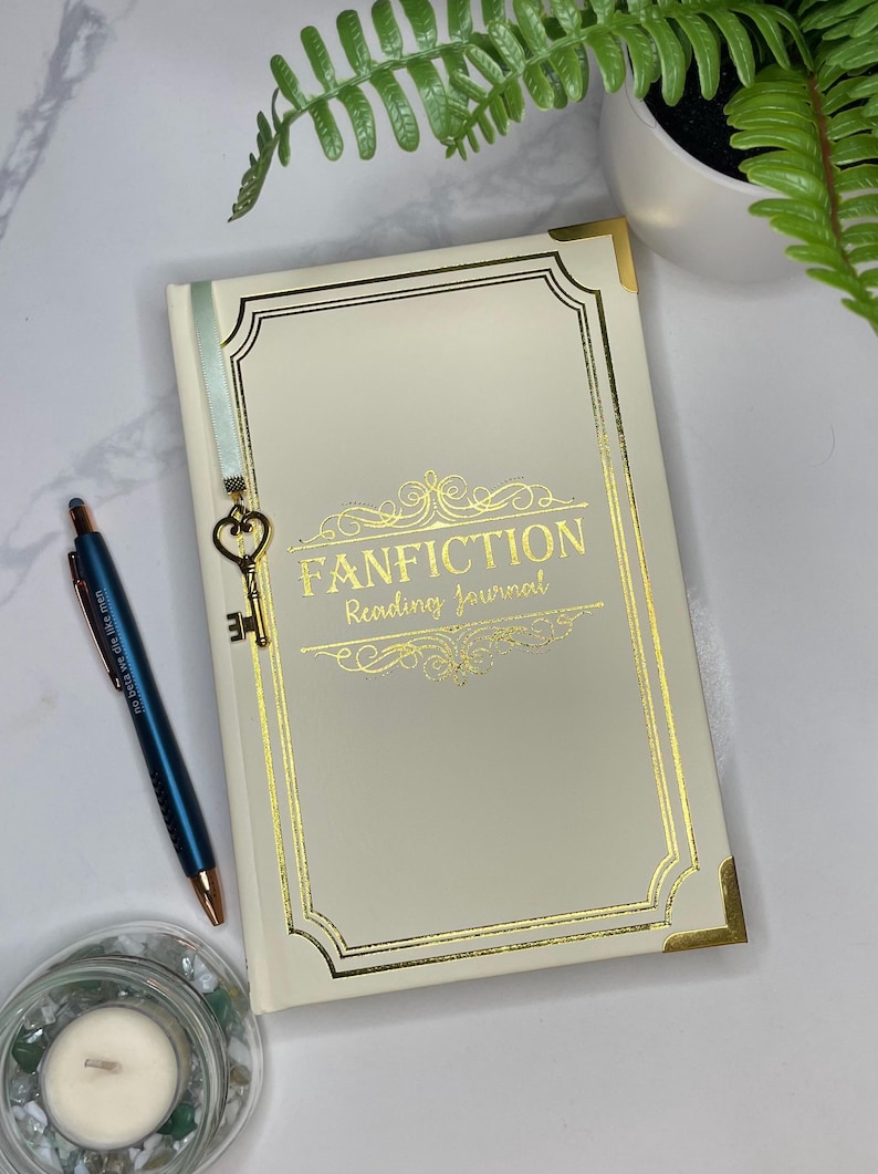 Peut inclure: Un journal blanc avec des accents dor&eacute;s et le texte "FANFICTION Reading Journal" sur la couverture. Un charme de cl&eacute; dor&eacute;e est attach&eacute; &agrave; un marque-page en ruban bleu.