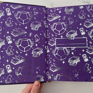 May include: A purple journal with a silver foil design featuring cats, stars, books, and hourglasses. The cover has a blank space for writing "To:" and a line for writing a name.