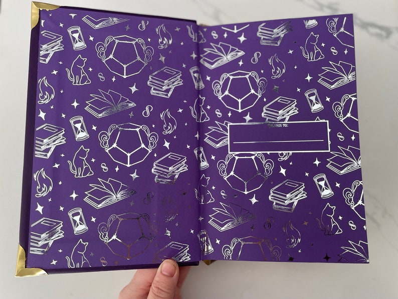 May include: A purple journal with a silver foil design featuring cats, stars, books, and hourglasses. The cover has a blank space for writing "To:" and a line for writing a name.