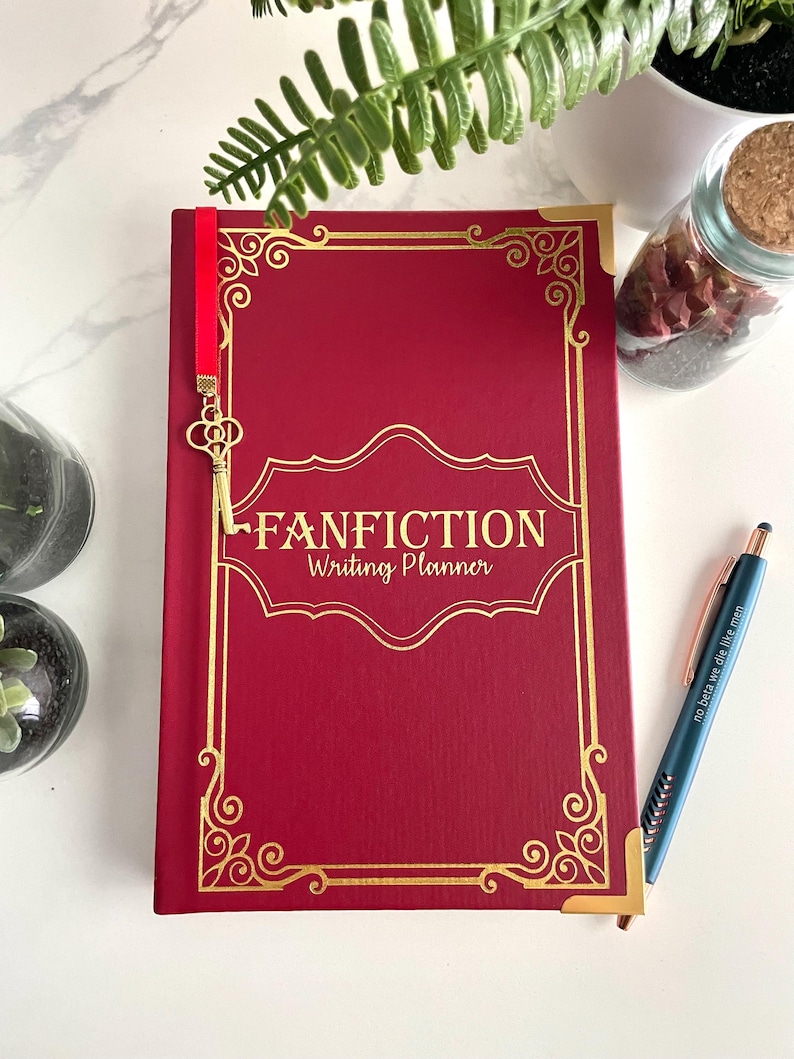 Pode incluir: Um di&aacute;rio de capa dura vermelho com detalhes dourados e um marcador de p&aacute;gina de chave dourada. O di&aacute;rio tem o texto "FANFICTION Writing Planner" na capa.