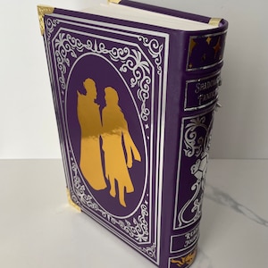 May include: A purple hardcover book with gold accents and a silver design. The book features a silhouette of two people in gold on the front cover. The title "Shadow and Bone" is visible on the spine. The book also has a "Reading Journal" label on the bottom.