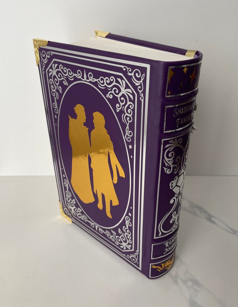 May include: A purple hardcover book with gold accents and a silver design. The book features a silhouette of two people in gold on the front cover. The title "Shadow and Bone" is visible on the spine. The book also has a "Reading Journal" label on the bottom.