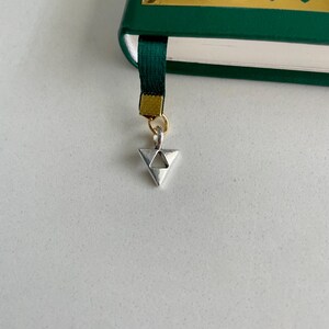 May include: A green book with gold accents and a dark green ribbon bookmark. The bookmark has a silver triangle charm attached with a gold ring. The book is on a white surface.