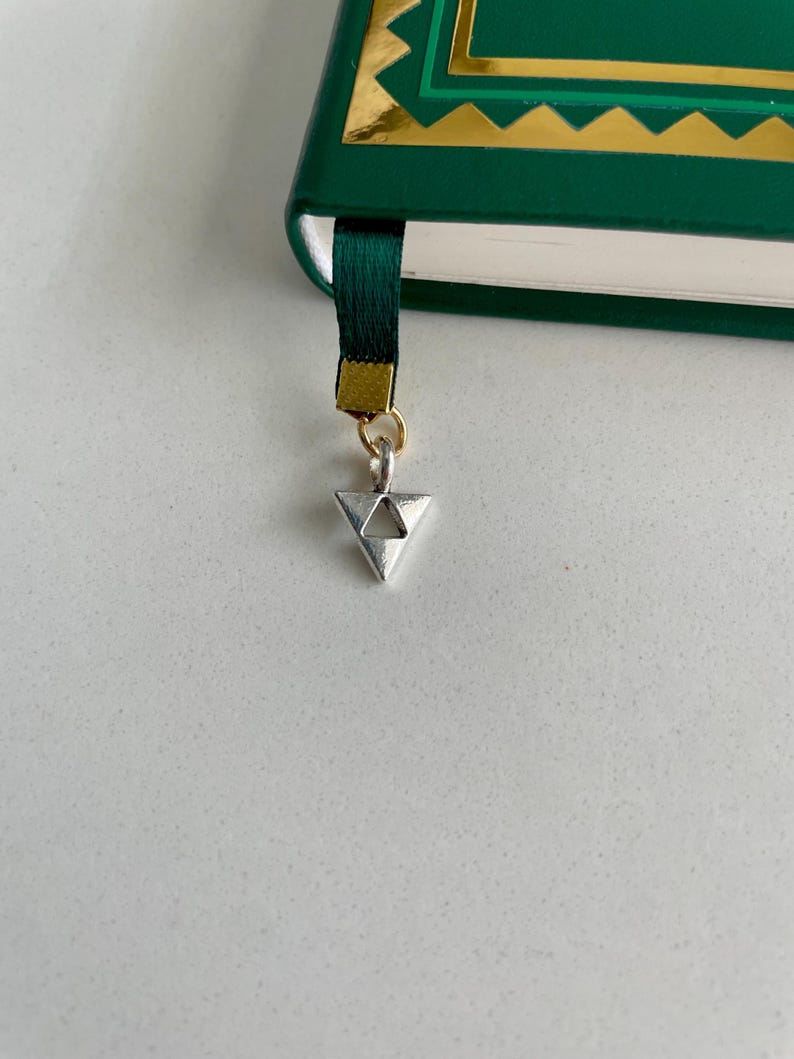May include: A green book with gold accents and a dark green ribbon bookmark. The bookmark has a silver triangle charm attached with a gold ring. The book is on a white surface.