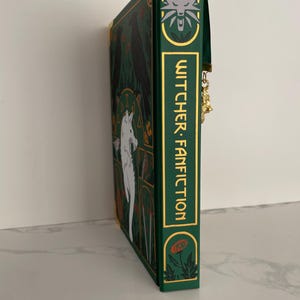 May include: A green book with a gold spine and a white wolf illustration on the cover. The book title is "Witcher Fanfiction" in gold lettering. The book has a green ribbon bookmark.
