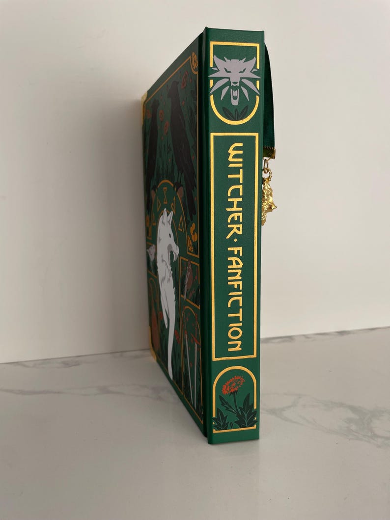 May include: A green book with a gold spine and a white wolf illustration on the cover. The book title is "Witcher Fanfiction" in gold lettering. The book has a green ribbon bookmark.