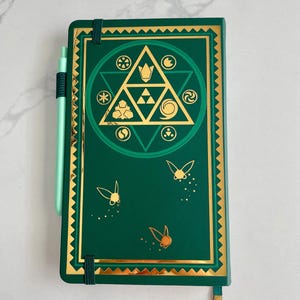 May include: A green notebook with gold accents and a matching pen. The cover features a gold geometric design and three gold butterfly illustrations. A gold bookmark hangs from the bottom. The notebook is closed with a green elastic band.