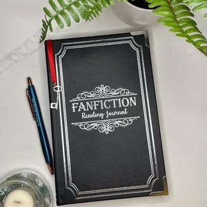 Peut inclure: Un journal noir avec des accents argent&eacute;s et le texte "FANFICTION Reading Journal" sur la couverture. Un ruban rouge est attach&eacute; au journal avec un charme de cl&eacute; argent&eacute;e.