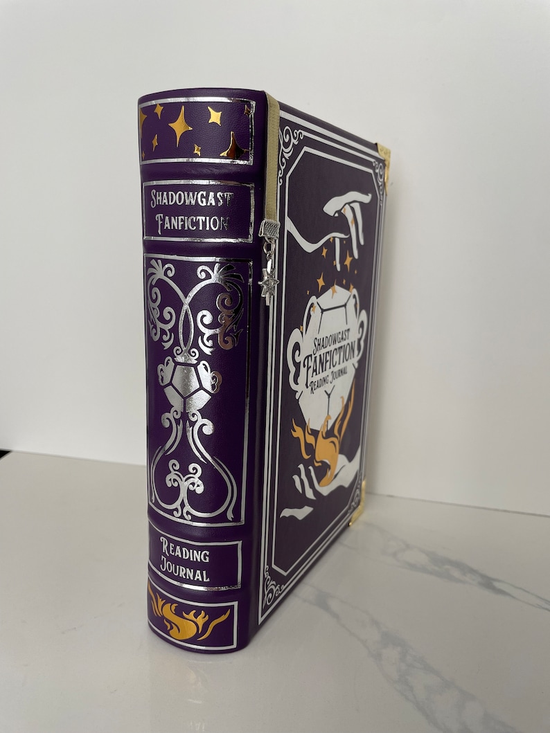 May include: A purple leather-bound journal with gold and silver accents. The cover features a hand holding a glowing orb and the text "Shadowgast Fanfiction Reading Journal".