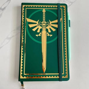 May include: Green notebook with a gold-colored sword and emblem design. The notebook has a gold border and a green elastic band. A light green pen is on the right side of the notebook. A silver charm hangs from the bookmark.