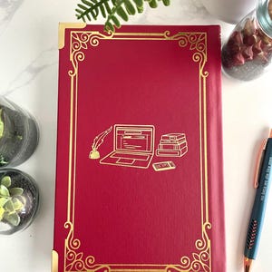 Pode incluir: Di&aacute;rio de capa dura vermelho com borda dourada e um design de um laptop, uma pena e uma pilha de livros. O di&aacute;rio tem as palavras "no beta we die like men" na lombada.
