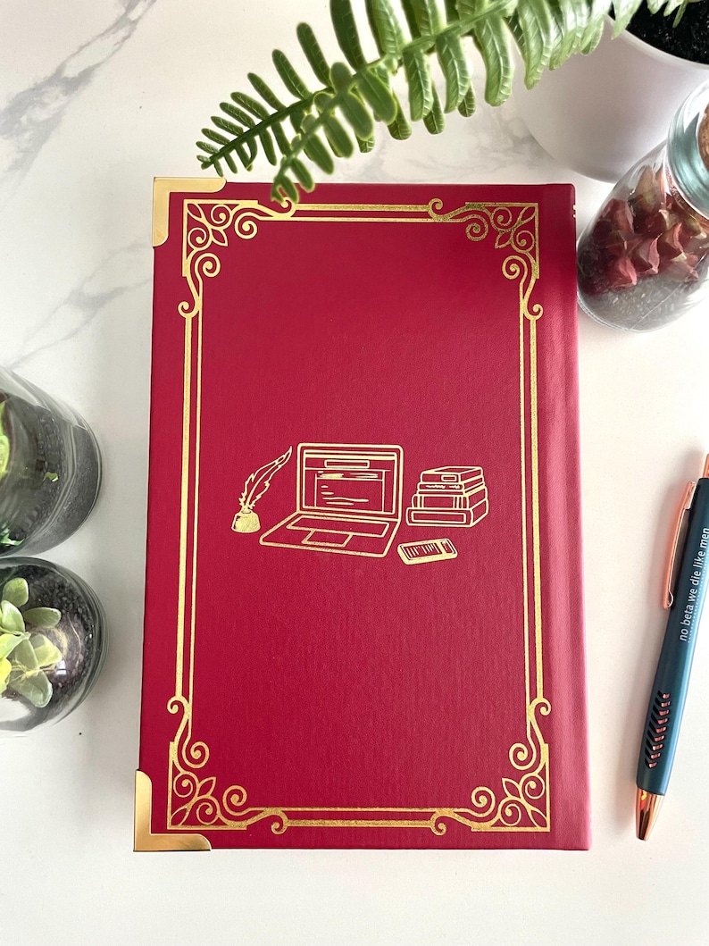 Pode incluir: Di&aacute;rio de capa dura vermelho com borda dourada e um design de um laptop, uma pena e uma pilha de livros. O di&aacute;rio tem as palavras "no beta we die like men" na lombada.