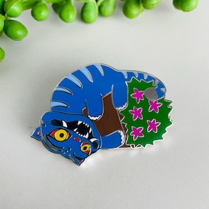 Puede incluir: Un pin de esmalte de gato azul con ojos amarillos y una sonrisa con dientes blancos. El gato está acostado junto a un arbusto verde con flores rosas. El pin tiene un borde plateado y está sobre un fondo blanco.