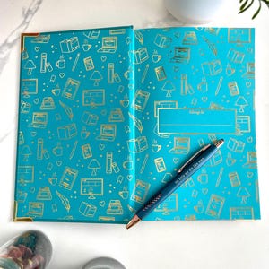 Peut inclure: Un carnet bleu turquoise avec des accents dor&eacute;s et un motif dor&eacute; de livres, tasses &agrave; caf&eacute; et autres objets li&eacute;s &agrave; la lecture. Le carnet a un espace vide pour un nom avec les mots "appartient &agrave;" au-dessus. Un stylo bleu avec les mots "so be it, we die like men" est pos&eacute; sur le carnet.