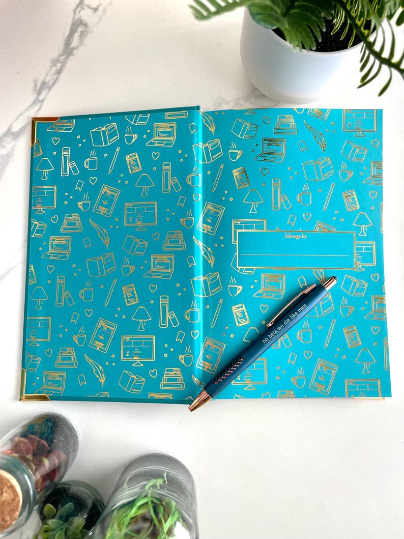 Peut inclure: Un carnet bleu turquoise avec des accents dor&eacute;s et un motif dor&eacute; de livres, tasses &agrave; caf&eacute; et autres objets li&eacute;s &agrave; la lecture. Le carnet a un espace vide pour un nom avec les mots "appartient &agrave;" au-dessus. Un stylo bleu avec les mots "so be it, we die like men" est pos&eacute; sur le carnet.