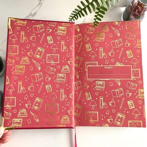 Pode incluir: Di&aacute;rio de capa dura vermelha com um design em folha de ouro de livros, x&iacute;caras de caf&eacute;, l&acirc;mpadas e computadores. O di&aacute;rio tem um espa&ccedil;o em branco na contracapa que diz "pertence a".