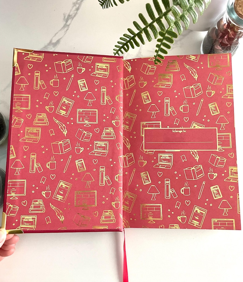 Pode incluir: Di&aacute;rio de capa dura vermelha com um design em folha de ouro de livros, x&iacute;caras de caf&eacute;, l&acirc;mpadas e computadores. O di&aacute;rio tem um espa&ccedil;o em branco na contracapa que diz "pertence a".