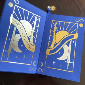 May include: A blue hardcover book with gold foil accents. The cover features a sun, moon, and two swords. The title "IMMORTAL HUSBANDS" is written in gold foil on the cover.