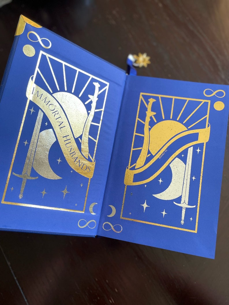 May include: A blue hardcover book with gold foil accents. The cover features a sun, moon, and two swords. The title "IMMORTAL HUSBANDS" is written in gold foil on the cover.