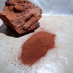 May include: A chunk of reddish-brown brick sits beside a pile of fine, reddish-brown powder. The brick has a textured surface with visible imperfections. The powder appears to be the result of the brick being ground down.