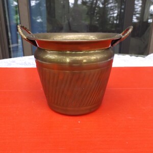 May include: A small, brass planter with two handles. The planter has a ribbed design and is a golden color.