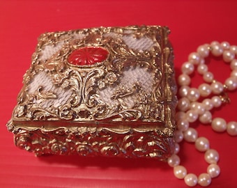 May include: A gold-colored ornate jewelry box with a red center piece and a pearl necklace on a red background.