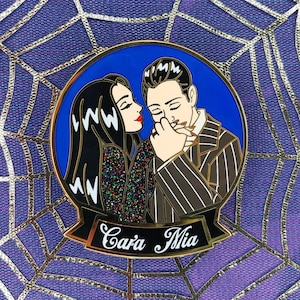 May include: A gold-colored enamel pin featuring a cartoon couple with black hair. The woman is wearing a sparkly black dress and the man is wearing a striped suit. The pin has a blue background and the text "Cara Mia" in a gold banner.