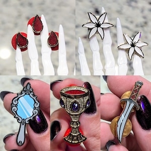 Pode incluir: Conjunto de pinos de esmalte com uma variedade de designs, incluindo uma lágrima vermelha, uma flor branca, um cálice dourado com gemas roxas, um espelho com um reflexo azul e uma adaga prateada com uma alça dourada.