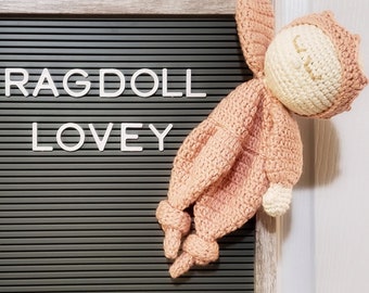 Puede incluir: Un muñeco de trapo de crochet con un cuerpo de color melocotón y una cara de color crema. El muñeco está colgado en un tablero de letras gris con las palabras "RAGDOLL LOVEY" escritas en letras blancas.