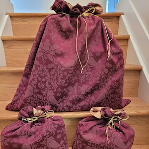 May include: Three burgundy fabric gift bags with gold ribbon ties. The bags feature a repeating pattern of birds, holly, and snowflakes.