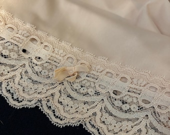 May include: A beige slip garment featuring a wide, intricate lace trim along the hemline. The lace displays a scalloped edge and a repeating floral pattern. A small, matching beige ribbon bow is centered on the lace trim, adding detail.