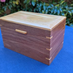 May include: A rectangular wooden box with a light-coloured top and a darker, reddish-brown body. The box features a small, light-coloured handle and decorative corner joints. The box is sitting on a blue surface.
