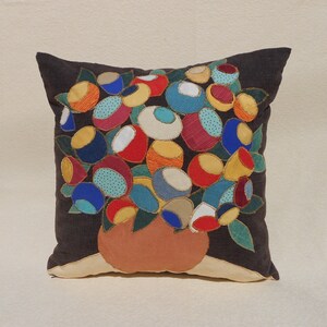 May include: A square dark brown throw pillow featuring an appliqued design of a terracotta-colored vase filled with a dense bouquet of rounded, multi-colored fabric shapes in shades of blue, red, yellow, orange, and white with gold stitching.