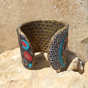 K&ouml;nnte beinhalten: Ein breiter Armreif mit einer texturierten goldenen Innenseite in Gitteroptik. Die Au&szlig;enseite zeigt ein mehrfarbiges Mosaikdesign mit schwarzen, roten und t&uuml;rkisfarbenen kreisf&ouml;rmigen Motiven sowie kleinen wei&szlig;en und schwarzen ringf&ouml;rmigen Mustern.