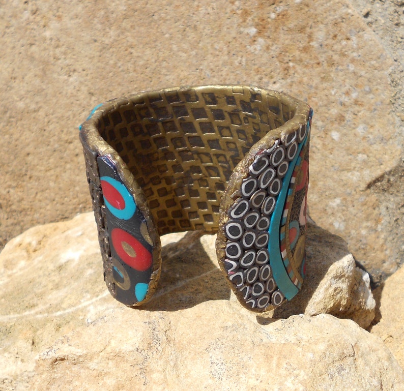 K&ouml;nnte beinhalten: Ein breiter Armreif mit einer texturierten goldenen Innenseite in Gitteroptik. Die Au&szlig;enseite zeigt ein mehrfarbiges Mosaikdesign mit schwarzen, roten und t&uuml;rkisfarbenen kreisf&ouml;rmigen Motiven sowie kleinen wei&szlig;en und schwarzen ringf&ouml;rmigen Mustern.