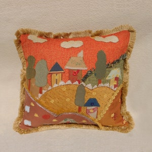 May include: A square decorative pillow featuring a textured orange background with a landscape scene made of fabric appliques. The scene includes houses, trees, and rolling hills in shades of green, yellow, and brown, finished with a tan fringe border.