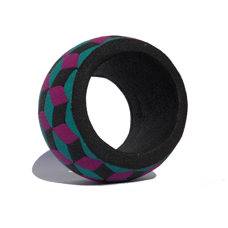K&ouml;nnte beinhalten: Ein breiter, kreisf&ouml;rmiger Armreif mit einer texturierten, matten Oberfl&auml;che. Die Au&szlig;enseite zeigt ein sich wiederholendes geometrisches Muster aus ineinandergreifenden W&uuml;rfeln in leuchtendem Magenta, T&uuml;rkis und Schwarz, w&auml;hrend das Innere schwarz ist.